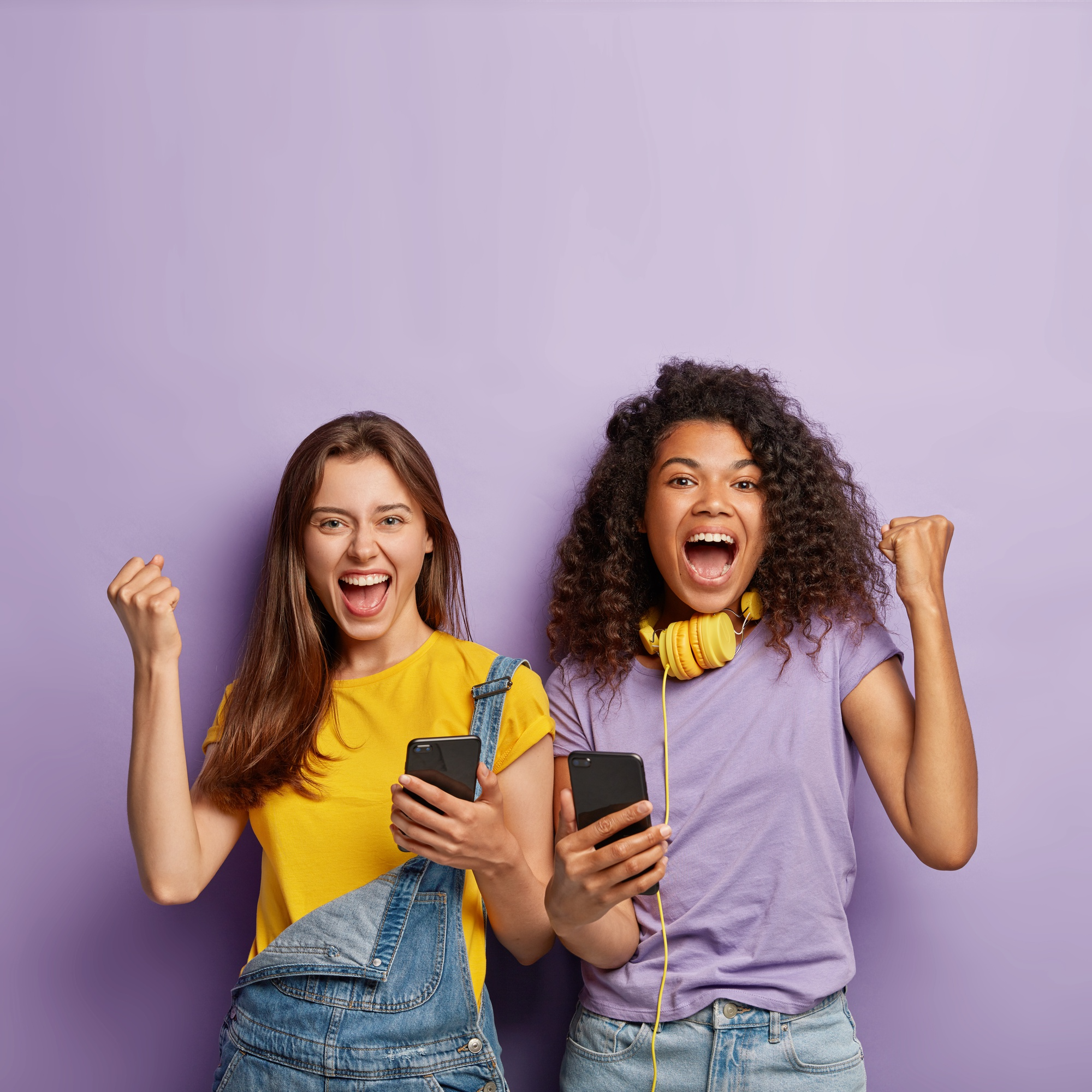 Two excited young women holding phones and celebrating, standing in front of a purple wall.